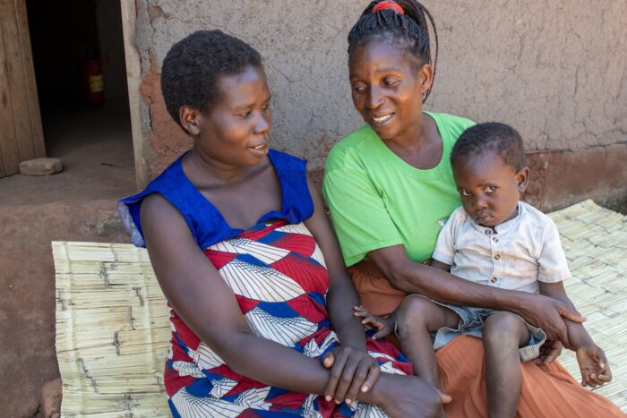 Mentor Mother holds a young boy as she talks to his mother while sitting on a woven mat.