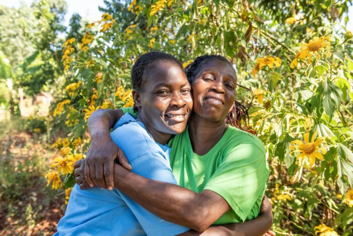 Two mentor mothers embrace in front of a bush covered in flowers.